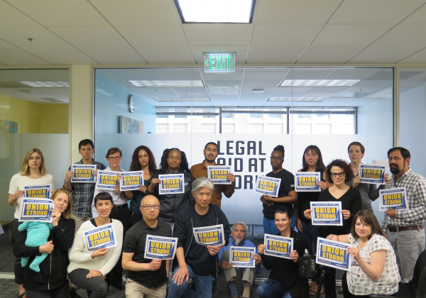Image of abut 25 Legal Aid at Work Staff standing in from of LAAW logo and holding signs that say "union strong"
