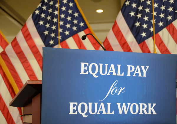 Sign saying "equal pay for equal work" in front of a podium, behind which sit two American flags