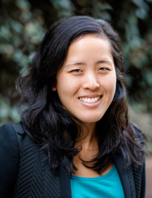 Headshot of Alison Kaneko smiling wearing a suit coat over a teal shirt in front of a leafy background
