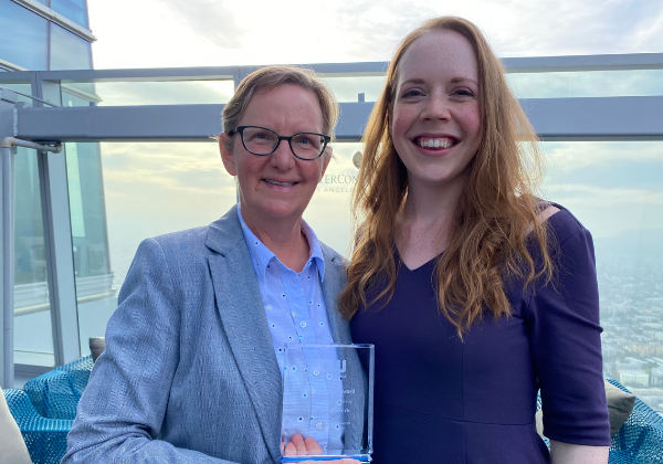 Attorneys Elizabeth Kristen and Katie Wutchiett standing with a clear plaque award on the deck of a large skyscraper with clouds in the background
