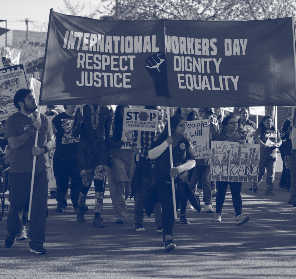 Group of people marching at a demonstration with banners and signs calling for "Respect, Justice, Dignity, and Equality" and "Stop Mass Incarceration & Deportations" in monochrome