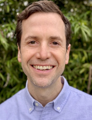 headshot of white male with brown hair and a blue button-down shirt smiling and standing in front of grenery