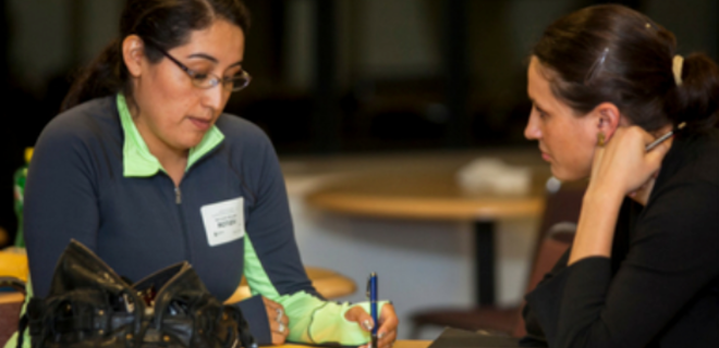 Two women sit at a table engaged in a serious conversation; one woman takes notes while the other listens attentively with her hand on her chin.