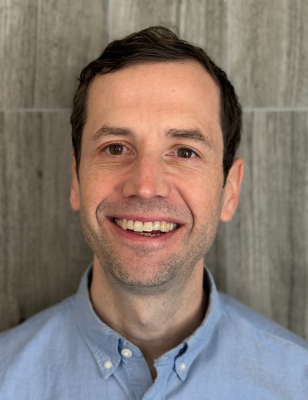 Headshot of a smiling man wearing a light blue button-down shirt against a neutral gray background.